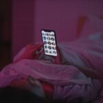 A woman relaxes in bed at night, browsing on her smartphone under pink ambient lighting.